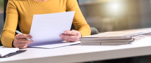 personne lisant et signant un document administratif sur un bureau, avec une pile de dossiers pour le dépôt de permis.