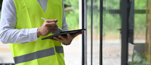 Etude thermique permis de construire : technicien en gilet jaune contrôlant une baie vitrée et notant des mesures sur une tablette.